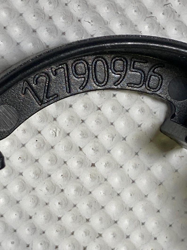 May include: Close-up of a black, curved plastic component with the number "12790956" embossed on its surface. The component has a matte finish and is set against a textured, white background.