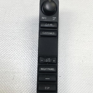 Puede incluir: Panel de control interior de coche negro con una perilla redonda en la parte superior y varios botones. Los botones están etiquetados como "INFO", "CLEAR", "CUSTOMIZE", "NIGHTPANEL", "ESP" y otros con símbolos.