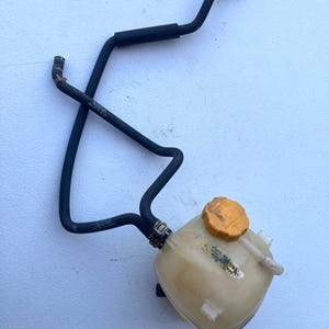 May include: A translucent white plastic coolant expansion tank featuring a yellow screw-on cap and two black rubber hoses attached with metal clamps. The hoses are shaped with bends and include small white plastic connectors at the ends.