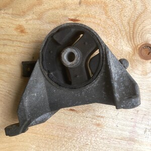 May include: A used, gray metal automotive engine mount with a black rubber insert. The mount has a curved shape with a central hole and several mounting points. The object is set against a wooden background.
