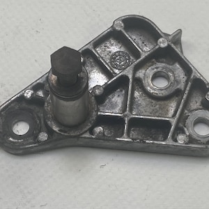 Puede incluir: Un soporte metálico plateado usado con un perno y un componente cilíndrico. El soporte tiene forma triangular con múltiples agujeros y una superficie texturizada. El perno es oscuro y hexagonal.