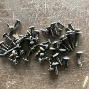 May include: A collection of small, dark gray screws with Phillips head and flat or rounded tops. The screws are scattered on a textured, brown surface, showing their threaded shafts.