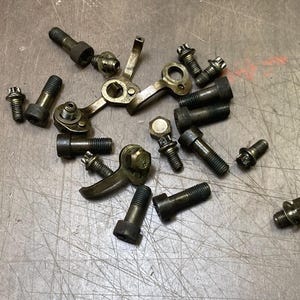 Peut inclure: Un assortiment de boulons, vis et composants mécaniques métalliques vintage de différentes tailles et formes. La quincaillerie est un mélange de tons bronze foncé et argent, dispersés sur une surface texturée gris clair.