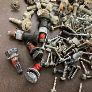 May include: A close-up of a variety of screws, bolts, and nuts in silver, black, and red. The screws are scattered on a brown surface.