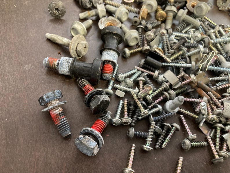 May include: A close-up of a variety of screws, bolts, and nuts in silver, black, and red. The screws are scattered on a brown surface.