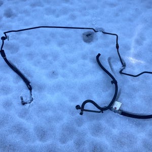 May include: A black automotive hose assembly, with various bends and fittings, lies on a snow-covered surface. The hose appears to be made of rubber and metal components, with a few small tags attached.