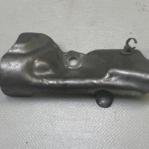 May include: A metallic gray automotive part, possibly a heat shield or exhaust component. It has a curved shape with a circular opening and a small clip. The surface shows signs of wear.