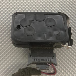 May include: A black automotive sensor with a rectangular shape and a silver metal clip. The sensor has a series of circular indentations on the top surface.