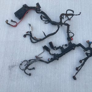 Peut inclure: Un faisceau de câblage électrique automobile noir composé de multiples câbles regroupés, de divers connecteurs en plastique, d'une boîte à fusibles noire et d'un fil rouge proéminent. L'ensemble comprend plusieurs supports de montage en métal.