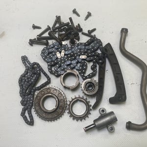 May include: An assortment of automotive engine parts, including chains, gears, bolts, and a curved metal pipe. The components are in various shades of black, silver, and gray, suggesting they are made of metal. The parts are scattered on a light-colored surface.