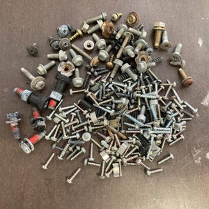 Puede incluir: Una gran colección de tornillos, pernos y tuercas surtidos en tonos plateados, negros y dorados. El hardware está esparcido sobre una superficie marrón.