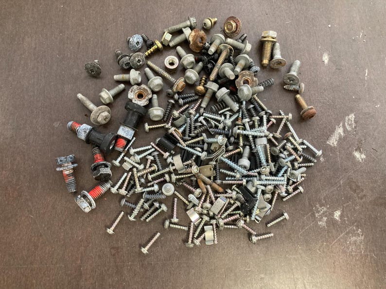 May include: A large collection of assorted screws, bolts, and nuts in silver, black, and gold tones. The hardware is scattered on a brown surface.