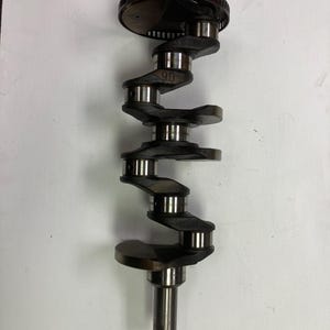 May include: A black metal crankshaft with a silver gear on the top. The crankshaft has six connecting rods and a long shaft.