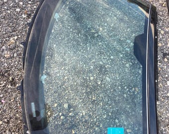 May include: A black car window with a curved top and a silver trim. The glass is clear, and a blue sticker is visible on the lower right corner. The window is lying on a gravel surface.