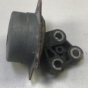 May include: Two automotive parts are shown. One is a dark gray, cylindrical rubber mount with a metal flange. The other is a dark gray, irregularly shaped metal bracket with three circular holes. Both parts appear used.