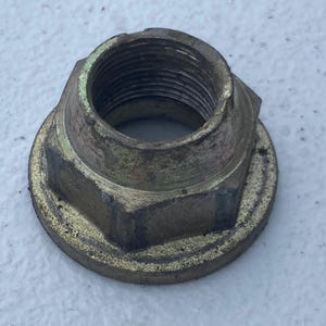 May include: A metallic, hexagonal flange nut with a weathered, brass-toned finish. The fastener features a wide, circular base and a central, threaded hole. The exterior surface shows signs of wear, including dark spots and surface oxidation.