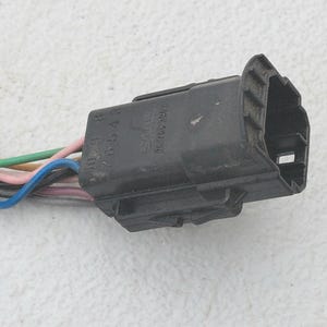May include: A black plastic automotive electrical connector with a multi-pin housing. Several insulated wires in colors including pink, green, blue, and grey extend from the base of the connector, which features molded text and numerical markings.