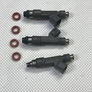 May include: Three dark gray fuel injectors with black connectors and copper-colored tips are displayed. Four red O-rings are also present. The components are arranged on a white, textured surface, suggesting automotive parts.