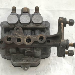 Op de afbeelding: Een grijze metalen brandstofpomp met vier inlaten en een zijdelings gemonteerde beugel. De pomp heeft een reeks gaten aan de bovenkant en een label met de tekst "62002-01-A111".