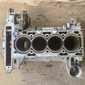 May include: A used, silver-colored engine block with four cylinder openings. The engine block has various attached components and is set against a wooden surface.