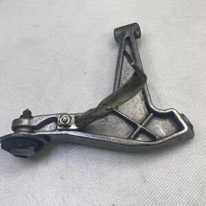 May include: A silver metal motorcycle foot peg with a black rubber foot pad. The foot peg is attached to a metal arm with a bolt.