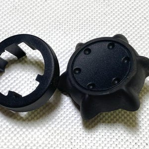 May include: Two black plastic components. One is a circular ring with cutouts, the other is a rounded shape with a flat top and six small circular indentations. The items are on a white textured surface.
