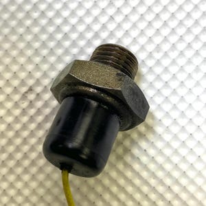 May include: A close-up of a mechanical component featuring a threaded metal end, a hexagonal nut, and a black cylindrical body. A yellow wire extends from the black body. The component appears to be a sensor or a similar device.