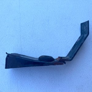 May include: A dark-colored metal bracket with an angled, L-shaped design. The component features a flat base, a central circular rubber grommet, and a bent arm extending upward with a small mounting hole at the top end. Signs of rust are visible.