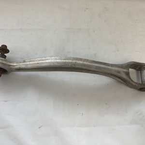 Op de afbeelding: Een zilveren metalen auto-onderdeel met een afgerond uiteinde en een lus aan het andere uiteinde. Het onderdeel heeft twee bouten die aan het afgeronde uiteinde zijn bevestigd.