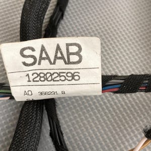 Peut inclure: Une étiquette blanche avec du texte noir qui indique "SAAB 12802596" et "AO 356231 B". L'étiquette est fixée à un faisceau de fils noirs, gris, bleus et rouges.
