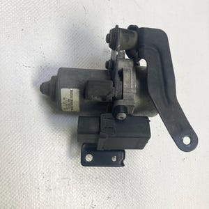 Op de afbeelding: Een auto-onderdeel, waarschijnlijk een motor, met een cilindrisch lichaam en bevestigde zwarte componenten. Het onderdeel heeft een label met tekst en staat tegen een witte achtergrond. De zwarte componenten omvatten een beugel en een rechthoekige connector.