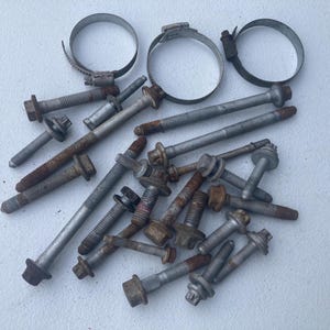 May include: A collection of assorted metal hardware including various steel bolts with hexagonal heads and threaded shafts, some showing signs of surface rust, alongside three circular metal hose clamps with adjustable screw mechanisms.