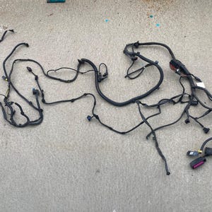 May include: A black automotive wiring harness featuring multiple branching cables, protective black conduit sleeves, and various plastic electrical connectors in colors including blue, white, and purple, arranged in a sprawling, complex configuration.