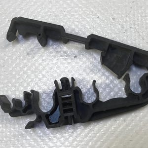 May include: A black plastic automotive part, likely a clip or bracket, with multiple curved and angular sections. The part appears to be new and unused, with a matte finish. The design includes several hooks and slots.