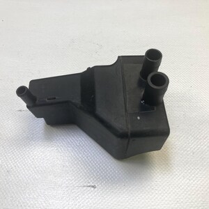 May include: A black plastic automotive part with two cylindrical tubes and one smaller tube extending from the main body. The part is rectangular with angled sides and sits on a white textured surface.