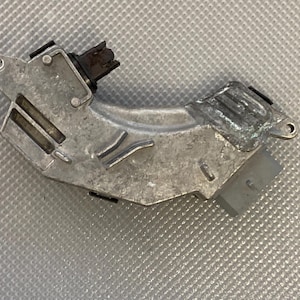 Puede incluir: Una pieza de automóvil de metal plateado con un conector negro y un cable plateado. La pieza tiene una forma curva y una serie de crestas en la superficie.