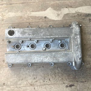 May include: A silver-colored engine valve cover with multiple circular openings and bolt holes. The cover has a rectangular shape with a curved section on one end. The metal surface shows signs of wear.