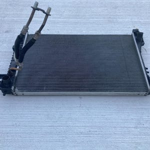 May include: A rectangular automotive radiator featuring a metallic core with horizontal cooling fins, black plastic side tanks, and a Y-shaped metal pipe assembly extending from the top left corner with two rubber hoses attached to the metal frame.