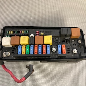 May include: A black fuse box with a variety of fuses and relays. The fuse box has a red wire attached to it.