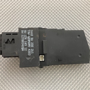 May include: A black electronic component with the text "SAAB KA 208 261" and other technical details. The component has a rectangular shape with various connectors and a textured surface. The background is a light gray, patterned surface.