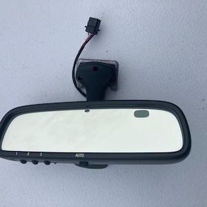 May include: A black automotive rearview mirror with an integrated mounting bracket and a short wiring harness with a connector. The mirror features three numbered buttons, an auto-dimming sensor, and the text AUTO printed on the lower frame.