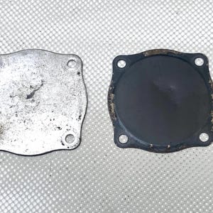 May include: Two metal plates with rounded corners and holes for screws. One plate is silver and worn, the other is black and circular. The plates are likely components of a mechanical device or engine.
