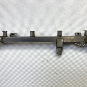 May include: A metallic silver fuel rail assembly with multiple cylindrical ports and fittings. The component has a series of connecting points and a curved pipe section. The item is likely an automotive part.