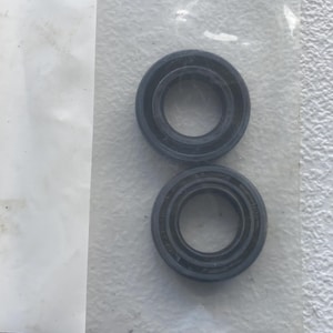 May include: Two circular black rubber oil seals with a smooth outer ring and a central opening. The components feature embossed text and markings on the surface, presented in a clear plastic packaging sleeve against a textured white background.