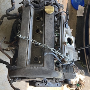 May include: A close-up of a gray car engine with black hoses and a chain attached to it. The engine is sitting on a concrete surface.