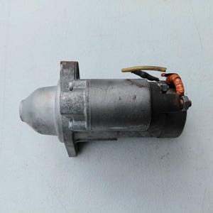 May include: A metallic automotive starter motor featuring a cylindrical housing, a mounting flange, and an electrical solenoid terminal with a short beige wire and a small orange-wrapped wire connection attached to the rear end.