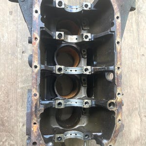 May include: A used, dark gray engine block with four cylindrical openings. The block has several bolt holes and metal bearing caps. The metal has some rust and wear.