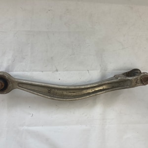 Op de afbeelding: Een zilveren metalen auto-ophangingarm met een rond uiteinde en een letter "R" gestempeld aan de zijkant.