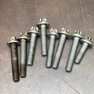 May include: A collection of eight silver-colored bolts with hexagonal heads and threaded shafts. The bolts are arranged on a brown surface, with varying degrees of wear and oxidation visible on the metal.