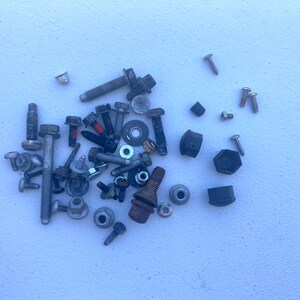 May include: A collection of assorted metal hardware including various bolts, screws, nuts, and washers of different sizes and finishes. The assortment features silver, grey, and dark metallic tones with some components showing signs of oxidation.
